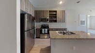 This single-floor unit offers a thoughtfully designed layout with a seamless flow between living, dining, and kitchen spaces. The kitchen is a standout feature, showcasing rich wood-tone cabinetry with open shelving, granite-style countertops, stainless steel appliances including a refrigerator, electric range, and microwave, and light hardwood-style flooring that extends into the main living area. The open kitchen layout includes a convenient peninsula, ideal for casual dining or meal prep.

The living area is bright and open, with large windows fitted with blinds that invite natural light while maintaining privacy. The space transitions smoothly into the private rooms, where plush carpeted flooring adds warmth and comfort. Each bedroom is generously sized, featuring ceiling fans with light fixtures and windows with views of surrounding greenery.

The bathroom is well-appointed with a wood-toned vanity, granite-style countertop, a large mirror, and a full tub-shower combination accented by tile surround. A tall storage cabinet provides additional organizational space. Clean white walls and light flooring throughout the unit create a neutral, airy ambiance, making the space highly adaptable to a variety of interior styles. The overall design balances modern finishes with functional everyday living.