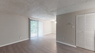 This single-floor unit offers a clean, functional layout with a neutral color palette throughout. The open living and dining area is finished with durable wood-look laminate flooring and features a textured popcorn ceiling. Vertical blinds adorn a full-length sliding glass door that floods the space with natural light and provides direct views of the surrounding greenery. A ceiling fan with light fixture serves the adjoining dining area, contributing to the airy feel of the combined living space. A wall-mounted thermostat is conveniently positioned for climate control.

The kitchen is efficiently designed with ample white cabinetry providing generous storage, light-toned laminate flooring, and white countertops with a double-basin sink. Black appliances — including a gas range, dishwasher, and refrigerator — offer a bold contrast to the bright cabinetry and walls, creating a practical and visually defined cooking space.

The bathroom is straightforward and functional, featuring a single vanity with a large mirror, a tiled tub-shower combo with a curved curtain rod, and white fixtures throughout. The overall ambiance of the unit is bright, clean, and move-in ready, with consistent flooring and neutral tones creating a cohesive feel across all areas.