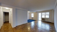 This single-floor space offers an open, airy layout that seamlessly connects its living areas. The main area is finished with classic parquet flooring arranged in a checkerboard pattern, lending a timeless character to the space. A partial wall divides the interior, creating a subtle sense of separation between zones while maintaining an open feel throughout.

The inner section of the space features blue carpeting, providing a visual contrast to the parquet and defining a distinct functional area. Two windows allow natural light to filter in from multiple directions, brightening the interior. A glass door leads to what appears to be an exterior balcony or outdoor area, further enhancing the sense of openness and light.

Climate control is provided by a wall-mounted PTAC unit positioned near the glass door, alongside a radiator unit visible beneath one of the windows. The walls are finished in a clean, neutral white and cream palette, offering a versatile backdrop. The layout is straightforward and practical, with clear sightlines across the space, making it well-suited for a variety of furnishing arrangements.