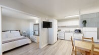 This single-floor unit offers a thoughtfully organized layout with a bright, neutral aesthetic defined by soft white walls and light hardwood-style flooring that runs consistently throughout the space. The bedroom provides a comfortable retreat, furnished with a full-size bed flanked by matching nightstands, a wall-mounted air conditioning unit, and a window with blinds and drape panels that allow for natural light and outdoor views of surrounding greenery. A large mirror and a multi-drawer dresser enhance the room's functionality and sense of space. Adjacent to the bedroom, the open kitchen is efficiently designed with an abundance of white upper and lower cabinetry, a skylight that draws in additional natural light, and a full suite of white appliances including a gas range, dishwasher, full-size refrigerator, and countertop microwave. A double-basin sink and ample counter space provide practical utility. A wall-mounted flat-screen television is positioned at the transition between the bedroom and the main living area, with a small dining area nearby featuring a wooden table and chairs. The bathroom is clean and straightforward, offering a tub-shower combination with tile surround, a single vanity with a speckled countertop, and a large mirror above. The overall ambiance is simple, functional, and well-maintained, suited for comfortable everyday living.