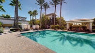 The pool area at Serenade at Riverpark in Oxnard, California offers a resort-inspired outdoor retreat designed for relaxation and recreation. At the heart of the space sits a generously sized, sparkling turquoise swimming pool with a stone-paved surround, framed by a striking canopy of towering palm trees that lend the area a classic Southern California ambiance. A separate spa with an accessible entry lift is tucked nearby, providing an additional amenity within the well-appointed grounds. Shaded cabanas and cushioned lounge chairs are arranged along the pool deck, offering comfortable spots to unwind under the abundant natural sunlight. A covered gazebo with outdoor seating provides a sheltered gathering area overlooking the pool. An outdoor dining patio features round tables with vibrant red chairs and large cantilever umbrellas, ideal for al fresco dining and socializing. Lush, meticulously maintained landscaping weaves throughout the entire area, including manicured hedges, flowering plants, ornamental shrubs, and mature trees. A curved architectural feature with floor-to-ceiling windows opens to a beautifully curated garden seating area, adding an elegant focal point to the communal grounds. Brick-paver walkways connect each zone seamlessly, completing this thoughtfully designed outdoor amenity space.