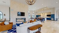 Located at 1196 Portside Dr in Ventura, CA, this single-floor community clubhouse is a thoughtfully designed gathering space that blends coastal elegance with modern comfort. The expansive open floor plan is anchored by soaring ceilings that amplify the sense of volume throughout, while an abundance of dramatic arched windows with black steel frames invites generous natural light and views of the surrounding community.

A striking floor-to-ceiling stone tile accent wall serves as the visual centerpiece of the main lounge, featuring a wide, linear gas fireplace set within a sleek black frame. Above it, a large wall-mounted TV enhances the entertainment-ready layout. A rustic orb chandelier hangs overhead, complementing the warm-toned wood and blue upholstered furnishings arranged on a bold patterned area rug.

The space transitions seamlessly into a dining and kitchenette area outfitted with navy blue cabinetry, white quartz countertops, stainless steel appliances, and a textured stone backsplash. Pendant lighting and a mounted TV complete this functional zone. Additional seating vignettes with lounge chairs, bar-height tables, and casual groupings are distributed throughout, offering flexible arrangements for both social gatherings and quiet retreats. Light hardwood flooring unifies the entire space.