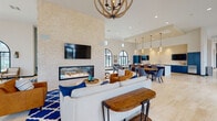 Located at 1196 Portside Dr in Ventura, CA, this single-floor community clubhouse is a thoughtfully designed gathering space that blends coastal elegance with modern comfort. The expansive open floor plan is anchored by soaring ceilings that amplify the sense of volume throughout, while an abundance of dramatic arched windows with black steel frames invites generous natural light and views of the surrounding community.

A striking floor-to-ceiling stone tile accent wall serves as the visual centerpiece of the main lounge, featuring a wide, linear gas fireplace set within a sleek black frame. Above it, a large wall-mounted TV enhances the entertainment-ready layout. A rustic orb chandelier hangs overhead, complementing the warm-toned wood and blue upholstered furnishings arranged on a bold patterned area rug.

The space transitions seamlessly into a dining and kitchenette area outfitted with navy blue cabinetry, white quartz countertops, stainless steel appliances, and a textured stone backsplash. Pendant lighting and a mounted TV complete this functional zone. Additional seating vignettes with lounge chairs, bar-height tables, and casual groupings are distributed throughout, offering flexible arrangements for both social gatherings and quiet retreats. Light hardwood flooring unifies the entire space.