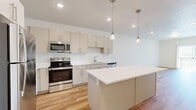 This single-floor, one-bedroom, one-bathroom unit offers a thoughtfully arranged layout that maximizes functionality within a compact footprint. The open-plan kitchen and living area serves as the heart of the unit, where warm-toned hardwood-style flooring runs throughout, creating a cohesive and inviting atmosphere. The kitchen is well-appointed with a full suite of stainless steel appliances, including a refrigerator, range, microwave, and dishwasher. Flat-front cabinetry in a neutral greige tone lines the walls and reaches toward the ceiling, offering generous storage. A central island with a white countertop provides additional workspace and is illuminated by two sleek glass pendant lights. Recessed lighting supplements the overall brightness of the space. The living area flows naturally toward a sliding glass door that opens onto a private balcony with mountain views. The bedroom features plush carpeting, a window with natural light, and direct access to a dedicated laundry closet housing a stacked washer and dryer. The bathroom is clean and modern, featuring a white vanity, a large mirror, and a full tub-shower combination with tile surround.