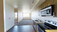 This 1-bedroom, 1-bathroom residence is designed with an open-concept layout that seamlessly connects the kitchen and living area. The kitchen features contemporary flat-front cabinetry in a warm wood-tone finish, stainless steel appliances including a range and built-in microwave, and a undermount sink set into a light stone-finish countertop. Track lighting runs along the ceiling, providing adjustable, focused illumination throughout the kitchen and adjoining space.

The transition from the kitchen leads into a spacious living area finished with plush carpeting, offering a comfortable and distinct zone for relaxing or entertaining. Large windows along the far wall allow natural light to fill the space, enhancing its open and airy feel. A secondary window is also visible near the entry corridor, further brightening the interior.

The layout includes a dedicated corridor leading to the bedroom and bathroom, maintaining a sense of separation between the living and private areas. Hard-surface flooring in the kitchen and entry contrasts with the carpeted living zone, adding visual interest and practicality to the overall design.