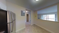 This single-floor unit presents a straightforward, functional layout with a clean and neutral aesthetic throughout. The main area features light-toned tile flooring and cream-painted walls, creating a bright and understated atmosphere. An electrical panel is mounted flush against the wall, offering practical accessibility. A window on the far wall allows natural light to filter in and provides an outward view of the surrounding building exterior, with a window-unit air conditioner installed beneath it for climate control.

A doorway leads into a connecting hallway finished with low-pile carpeting, which transitions the space toward additional rooms. The hallway features its own ceiling light fixture and opens to what appears to be a closet with a door at the far end, offering useful storage. A stainless steel refrigerator is partially visible along one wall of the main area, indicating proximity to a kitchen space.

The ceilings are smooth and of standard height, with flush-mount lighting providing even illumination throughout. White baseboards line the floors, adding a clean, finished look. Overall, the space is practical and move-in ready, with a neutral palette suited to a variety of uses.