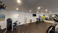 This dedicated fitness center is a fully outfitted single-level facility designed to support a wide range of workout routines. The space features durable rubber flooring throughout, providing a safe and functional surface for all types of exercise. Recessed lighting illuminates the room evenly, creating a bright and energizing atmosphere.

The cardio zone is well-stocked with multiple treadmills, elliptical trainers, a stationary recumbent bike, and a rowing machine, offering users diverse options for aerobic training. A wall-mounted flat-screen television adds convenience for those looking to stay entertained during workouts.

The strength training area includes a multi-function cable machine, an adjustable weight bench, a rack of dumbbells in varying sizes, and leg exercise equipment, making it suitable for full-body resistance training. Stability balls and additional accessories are also available for functional fitness exercises.

A large floor-to-ceiling mirror spans a significant portion of one wall, visually expanding the space and allowing users to monitor their form during workouts. Coastal-themed artwork adorns the walls, contributing a calm and inviting aesthetic. Louvered shuttered windows allow natural light to filter into the room. Two entry doors provide convenient access points, and the facility is equipped with standard safety signage.