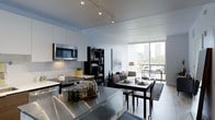 This contemporary single-floor space showcases a thoughtfully designed open-concept layout that seamlessly integrates the kitchen, dining, and living areas into one cohesive living environment. The kitchen is outfitted with crisp white cabinetry, a stainless steel countertop island, a built-in microwave, and a smooth cooktop, all illuminated by modern track lighting mounted on a clean white ceiling. The bright, neutral color palette throughout creates a sense of openness and visual continuity. A dark-toned bookcase adds a striking contrast and serves as a stylish divider between the kitchen and living zones. The dining area features a dark wood table paired with matching chairs, positioned beneath a large-scale art piece that anchors the space. The living area flows naturally toward expansive floor-to-ceiling windows that flood the interior with natural light and frame impressive views of the surrounding cityscape and greenery. Light wood-toned flooring runs throughout, complementing the modern furnishings. The overall ambiance is clean, urban, and sophisticated, making it an ideal space for both everyday living and entertaining.