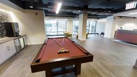 This single-floor amenity space is thoughtfully designed to blend recreation and relaxation in a contemporary setting. The centerpiece of the room is a full-size billiards table with a rich red felt surface and solid wood construction, offering an inviting focal point for social gatherings. The open-concept layout provides ample space around the table for comfortable gameplay.

Large floor-to-ceiling windows line the perimeter, flooding the interior with natural light and framing dynamic views of the surrounding urban streetscape. The industrial-style exposed ceiling features dark-painted ductwork and structural elements, complemented by sleek pendant and recessed lighting that adds a modern edge to the design.

The flooring throughout is a warm wood-toned plank, lending a clean and cohesive aesthetic to the space. Structural columns are finished in crisp white, reinforcing the contrast between the raw ceiling and the polished lower environment. A casual seating area positioned near the windows features upholstered lounge chairs in muted gold and burgundy tones, creating a comfortable retreat for conversation or quiet relaxation. The overall ambiance is modern and energetic, making this space well-suited for both informal socializing and community-oriented recreation.