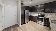 This thoughtfully designed 1-bedroom, 1-bathroom residence offers a cohesive single-floor layout that maximizes functionality and style. The open-concept kitchen is a standout feature, showcasing rich dark espresso cabinetry, stainless steel appliances including a refrigerator, range, and microwave, complemented by a decorative mosaic tile backsplash and granite countertops that extend to a breakfast bar peninsula. Pendant lighting and track lighting illuminate the space with a modern touch. Light wood-style plank flooring flows throughout the main living areas, creating a warm and contemporary aesthetic. The living area is open and airy, offering a clean canvas with neutral gray walls and smooth white baseboards. The bedroom features plush carpeting, natural light from a window fitted with blinds, and a calm, restful atmosphere. The bathroom is appointed with matching granite countertops, a dark vanity with an undermount sink, a large wall mirror, and tile flooring, combining practicality with refined design. The 3D dollhouse view reveals a well-organized floor plan with a clear, efficient flow between the living, sleeping, and bathroom areas, making this a practical and attractive residential unit.
