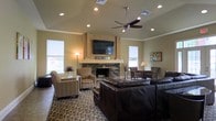 This single-story living space is defined by its vaulted ceiling, which adds a sense of openness and volume to the room. A ceiling fan with a brushed metal finish is centrally mounted, providing both function and visual balance overhead. Recessed lighting is distributed throughout, supplementing the warm ambient glow from floor and table lamps.

The focal point of the room is a stacked-stone fireplace framed by a tile surround and a light-toned mantel. A large flat-screen television is mounted directly above within a tile-accented housing, creating a unified entertainment and hearth feature. The neutral, warm-toned walls complement the natural textures of the stone and surrounding finishes.

Seating is generously arranged throughout the space, including a substantial dark leather sectional sofa and multiple patterned accent chairs that add visual interest and texture. A patterned area rug anchors the seating arrangement. Multiple windows dressed with roller shades allow natural light to filter in from several directions. A framed piece of colorful artwork decorates one wall, adding a subtle decorative accent to the overall warm and inviting atmosphere.