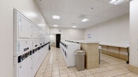 Located at 4710 Bethesda Ave in Bethesda, Maryland, this single-floor commercial laundry facility is designed for efficiency and user comfort. The space is finished with light beige tile flooring throughout and features a drop ceiling with recessed fluorescent panel lighting, creating a bright, clean environment.

The main laundry area is well-equipped with a substantial row of commercial-grade front-loading washers and top-loading machines, all offering digital controls for ease of use. Alongside the machines, a long bank of stacked dryer units lines one wall, providing extensive drying capacity. Central folding counters with a tiled base offer generous surface space for sorting and folding laundry, complemented by convenient waste receptacles and laundry bins positioned throughout the room.

A dedicated waiting area is furnished with modern chrome-framed upholstered chairs arranged for comfort, set against subtly striped wall paneling that adds a touch of texture to the otherwise neutral palette. Paper towel dispensers and informational signage are thoughtfully placed for patron convenience.

The layout is open and spacious, allowing for easy movement between machines and work surfaces. The facility's clean, functional design and well-maintained equipment make it a practical and organized laundry destination in the heart of Bethesda.