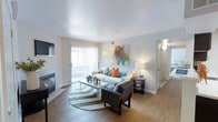 This thoughtfully designed single-floor residence offers a comfortable and inviting living environment filled with natural light. The main living area serves as the heart of the home, showcasing warm hardwood-style flooring, smooth white ceilings, and soft neutral walls that create a clean, airy backdrop. A fireplace anchors one side of the room, adding both warmth and a focal point to the space. Large sliding glass doors with vertical blinds open to a private balcony, allowing abundant natural light to flood the interior while offering a seamless connection to the outdoors. The living area is generously proportioned, providing ample room for comfortable seating arrangements centered around a stylish area rug. Vibrant accent pieces and colorful wall art add personality and visual interest to the otherwise understated palette. A side table with warm lamp lighting contributes to the cozy ambiance during evening hours. A hallway visible from the living area leads to additional rooms, suggesting a well-organized and functional floor plan. The overall atmosphere of this residence is bright, modern, and livable, with a balance of practical design and welcoming aesthetic touches throughout.