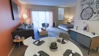 This well-appointed single-floor studio seamlessly integrates living, dining, and sleeping zones into one cohesive, open-plan layout. The dining area anchors the foreground with a round white table surrounded by modern seating, elegantly set with place settings and a decorative centerpiece. Beyond it, the living area features a plush sofa, a sleek accent chair, and a central coffee table set atop a patterned area rug, creating a comfortable and inviting lounge space. The sleeping area is thoughtfully positioned toward the rear of the room, offering a neatly made bed flanked by bedside lighting that adds a warm, restful ambiance. Large sliding glass doors stretch across the back wall, flooding the entire space with generous natural light and providing access to an outdoor area. Soft, neutral-toned carpeting runs throughout, complementing the warm wall colors and the light, airy curtains framing the doors and windows. The overall atmosphere is bright, modern, and harmoniously designed, with each zone flowing naturally into the next to make excellent use of the available space.