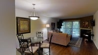 This single-floor space offers a welcoming and functional layout, highlighted by an open-concept design that connects the dining and living areas into one cohesive living environment. The rich, dark hardwood flooring runs throughout, grounding the space with warmth and a sense of continuity.

The dining area is anchored by a classic wooden table and matching chairs, illuminated by a pendant light fixture that adds a touch of elegance. A framed artwork on the wall completes the defined yet open dining nook.

The living area flows naturally from the dining space, centered around a plush, generously sized sofa accented with teal throw pillows. A dark wood coffee table and matching side table provide practical surfaces, while a table lamp adds soft, ambient lighting. Decorative wall art adds personality and visual interest to the room.

Large vertical blinds covering what appears to be a sliding glass door allow for natural light control and suggest access to an outdoor area, enhancing the overall brightness and airiness of the space. The warm, neutral wall tones complement the furnishings and flooring, creating a cohesive and inviting atmosphere throughout this well-appointed single-floor residence.