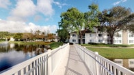 This single-floor property is situated within a beautifully landscaped waterfront community that evokes a calm, resort-like atmosphere. A standout feature of the setting is the white-railed pedestrian bridge that stretches alongside a peaceful pond, offering picturesque views of the still water and its natural reflections. Mature trees — including sweeping tropical palms and broad-canopied hardwoods — frame the surroundings, providing ample shade and a lush, green backdrop throughout the grounds. The community's exterior architecture features clean white stucco facades with a classic, understated Florida style that blends harmoniously with the natural environment. Well-maintained lawns extend to the water's edge, reinforcing the sense of openness and tranquility. The grounds include what appears to be a clubhouse or community facility visible across the water, hinting at shared amenities within the development. Natural light plays a prominent role in the outdoor ambiance, with bright Florida skies casting a warm, inviting glow across the walkways and water features. The overall character of this property and its surroundings conveys a peaceful, community-oriented lifestyle enriched by natural beauty and thoughtful landscaping.