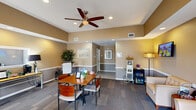 Located at 300 20th Ave W in Milan, Illinois, this single-floor office space serves as a well-appointed leasing and reception facility. The interior is finished in warm tan and gray tones with white chair-rail wainscoting, creating a professional yet inviting atmosphere throughout.

The main reception and lobby area is thoughtfully arranged with a central dining-style table and chairs for meetings or consultations, complemented by a comfortable seating arrangement featuring upholstered armchairs and a sofa. A wall-mounted flat-screen TV adds a modern amenity for waiting guests. A ceiling fan with integrated lighting anchors the tray ceiling overhead, while recessed lighting ensures the space is well-illuminated throughout. A beverage cooler and refreshment station add a hospitable touch for visitors.

The private office is equipped with a dedicated workstation, a secondary console desk, and natural light from multiple windows fitted with vertical blinds. A French door connects the office to the reception area, maintaining an open yet defined workspace. The tile and patterned carpet flooring transitions smoothly between areas, adding visual interest underfoot. A bathroom and additional utility spaces complete the layout, making this a fully functional and professionally presented office environment.