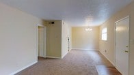 This single-story space presents an open layout connecting the living and dining areas through a straightforward, functional design. The dining area is anchored by a classic multi-arm chandelier that adds a traditional touch to the room, while a window along the adjacent wall allows natural light to filter in, brightening the space. Warm beige walls run throughout, complemented by white baseboards that provide clean contrast.

Wall-to-wall carpeting in a neutral tan tone covers the floors, contributing to a quiet, comfortable atmosphere. A textured ceiling spans the entirety of the space, adding subtle visual depth overhead. On the opposite end of the room, a doorway opens to what appears to be a hallway or closet area fitted with louvered doors, suggesting dedicated storage. A HVAC vent is visibly integrated into the ceiling near a structural column, indicating a centralized climate control system.

The overall layout is straightforward and uncluttered, offering a blank canvas with practical infrastructure already in place. The neutral color palette and traditional fixtures give the space a timeless, approachable character.