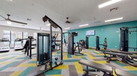 This modern fitness center is designed to provide a comprehensive workout experience within a spacious, single-floor layout. The interior is immediately distinguished by its bold teal accent walls, which lend a vibrant, energetic atmosphere throughout the space. Underfoot, colorful geometric-patterned carpet tiles in teal, yellow, and gray add a dynamic visual element, while designated zones transition to artificial turf, creating a dedicated functional training area.

The strength training section is thoroughly outfitted with a variety of cable machines, weight benches, a power rack, and a full rack of dumbbells, catering to a wide range of resistance training needs. Multiple flat-screen televisions mounted on the walls offer entertainment during workouts. Large wall-mounted mirrors extend across key areas of the room, enhancing the sense of openness and allowing users to monitor their form.

The cardio zone features a row of treadmills and an elliptical machine positioned along a wall of large windows, which flood the area with natural light and offer pleasant views of the surrounding greenery. Stability balls and additional free weights round out the functional fitness offerings. Ceiling fans and recessed fluorescent lighting ensure a comfortable, well-lit environment throughout. The overall ambiance is modern, motivating, and thoughtfully designed to support a full range of fitness activities.