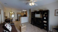 Located at 334 Carolina Forest Blvd in Jacksonville, NC, this single-story home offers a practical and welcoming open-concept design that seamlessly connects the main living areas. The living room is comfortably proportioned, featuring neutral carpeting underfoot and a ceiling fan with integrated lighting that keeps the space airy and well-lit. A large entertainment center serves as a natural focal point, providing ample storage and display space alongside a mounted television. The kitchen is positioned adjacent to the living room and is defined by a peninsula-style breakfast bar that creates a natural division between the two spaces while maintaining an open flow. The kitchen is equipped with light wood cabinetry, a stainless steel refrigerator, a microwave, and a dedicated workspace, all illuminated by pendant lighting overhead. A window in the kitchen area allows natural light to filter in, brightening the cooking space. The overall aesthetic throughout the home is neutral and versatile, with warm tones that lend a cozy, lived-in ambiance. The single-floor layout ensures ease of navigation and accessibility throughout the home, making it a functional and inviting residence in the Jacksonville, NC community.