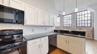 Located at 1287 West 9th Street in Cleveland's vibrant Ohio City neighborhood, this two-bedroom, two-bathroom unit blends industrial loft character with modern comfort. The open kitchen and living area anchors the space, showcasing exposed brick walls, soaring ceilings with ceiling fan, and warm hardwood flooring throughout. The kitchen is well-appointed with white shaker cabinetry, dark granite countertops, a classic subway tile backsplash, and black appliances including a range, dishwasher, and over-the-range microwave. Multi-pane industrial-style windows with red frames flood the main living area with natural light while reinforcing the building's historic character. The living room offers generous open space with continued hardwood floors and clean, light-toned walls. Both bedrooms are carpeted for comfort; the primary bedroom features an interior window overlooking the main living space, adding a distinctive loft feel. The two bathrooms are finished with wood-look tile flooring and full tub-shower combinations. Carpeted stairs lead to the unit entry, offering a welcoming transition into the space. Throughout, the interplay of brick, wood, and neutral tones creates a cohesive, urban-industrial ambiance.