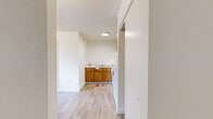 Located at 6502 E Golf Links Rd in Tucson, Arizona, this single-floor unit offers a practical and efficiently designed layout across a compact footprint. The interior features light wood-look plank flooring throughout, lending a clean and cohesive aesthetic to every room.

The kitchen is equipped with warm-toned wood cabinetry, a white electric range, and a full-size refrigerator, all set beneath a flush ceiling light. A built-in shelving nook adjacent to the kitchen provides convenient additional storage. The open layout allows the kitchen to flow naturally into the surrounding living space.

The main living area is bright and open, with neutral cream-toned walls and a textured ceiling. A vertical blind-covered window allows for natural light while maintaining privacy. A wall-mounted thermostat and ceiling vents indicate a central HVAC system for year-round climate control.

A separate room off the main living area features its own window and direct access to a bathroom, which includes a vanity with wood cabinetry and a mirror with overhead lighting. An additional utility area is visible, housing a water heater and storage space. White trim and clean lines throughout give the unit a simple, functional character well-suited to the Tucson climate.