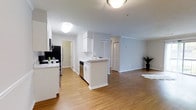 Located at 1235 Ashbourne Rd in Elkins Park, PA, this single-floor residence offers a clean, contemporary living environment designed for both comfort and functionality. The open-concept layout seamlessly connects the kitchen and living area, creating a sense of openness and ease of movement throughout the space. The kitchen is outfitted with white cabinetry, granite-style countertops, stainless steel appliances including a refrigerator and dishwasher, and a peninsula that serves as both a prep surface and a casual dining or gathering spot. Hardwood-style flooring runs throughout, adding warmth and visual continuity to the space. The living area is generously sized, providing ample room for various furniture arrangements, and is bathed in soft, ambient light from flush-mounted ceiling fixtures. Neutral gray and white tones on the walls lend the space a modern, move-in-ready aesthetic. Louvered closet doors offer practical storage solutions, while clean white baseboards and trim add a polished finishing touch. The layout also includes access to additional rooms via a hallway visible from the main living space, suggesting a well-organized and thoughtfully designed floor plan throughout.