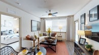 This single-floor residence offers a comfortable and thoughtfully designed living environment. The living area serves as the heart of the space, showcasing warm hardwood-style flooring, neutral toned walls, and a ceiling fan with integrated lighting that enhances both comfort and ambiance. Large windows dressed with layered blinds and patterned curtains allow abundant natural light to pour in, creating a bright and inviting atmosphere throughout the day. The open layout connects the living area seamlessly to a dining space, offering a practical and social-friendly flow. The living room is generously proportioned, providing ample room for a full sectional sofa accented with colorful throw pillows, a woven coffee table, and a vibrant area rug that adds warmth and texture underfoot. Decorative elements such as candlesticks, framed wall art, and carefully placed accent furniture contribute to a curated, homey aesthetic. The dining area accommodates a round table with place settings, positioned conveniently adjacent to the main living space. Overall, the residence strikes a balance between modern practicality and warm, welcoming design, making it a comfortable and functional single-level home.