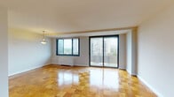This well-organized residential unit spans a single floor and is defined by its gleaming parquet wood flooring, which extends consistently throughout the living spaces and bedrooms, adding warmth and character to the interior. The spacious living and dining area is a standout feature, bathed in natural light through large sliding glass doors that open onto a private balcony overlooking a lush urban skyline. A side window further enhances the brightness of this generous main living space. The kitchen is clean and functional, featuring gray laminate countertops, a gas cooktop, white cabinetry, and classic black-and-white diamond-patterned tile flooring. The bedrooms are well-proportioned, each offering natural light through large windows with urban views, and are equipped with built-in closets featuring bifold doors and shelving. The primary bedroom benefits from an en-suite bathroom tiled in gray, complete with a bathtub. A second bathroom features retro mint-green tile and a matching tub, adding a charming vintage touch. Radiator heating units are present throughout, and the neutral white walls provide a versatile backdrop. The overall layout, as seen in the dollhouse view, is practical and spacious, making excellent use of the available floor plan.