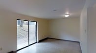 This two-floor space presents a clean, functional layout with straightforward finishes throughout. The pictured room features smooth, light-colored walls with a neutral palette that contributes to a bright and open atmosphere. A large sliding glass door dominates one wall, allowing abundant natural light to fill the interior while providing direct access to the outdoors. Through the glass, a sloped, landscaped area is visible, suggesting a ground-level or below-grade setting for this particular room. The flooring appears to be a speckled, light-toned material — consistent with a durable, low-maintenance surface suited for everyday use. A flush-mount ceiling light fixture provides overhead illumination, complementing the natural light from the sliding door. The walls are finished with simple baseboard trim, and electrical outlets are conveniently placed along the perimeter. The room's open layout offers flexibility in how the space can be arranged or utilized. With two floors available, the overall property provides ample room for a variety of functional configurations across its levels.