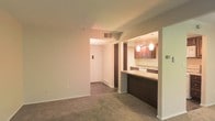 This two-floor residential space offers a practical and comfortable layout designed for everyday living. The main living area features neutral wall-to-wall carpeting that adds warmth underfoot, with smooth walls and a ceiling-mounted ventilation system providing a clean, understated aesthetic throughout.

A standout feature is the open pass-through kitchen, which connects seamlessly to the living space. The kitchen is appointed with rich dark wood cabinetry, light stone-toned countertops, and a built-in breakfast bar that doubles as an informal dining or workspace. Warm pendant lighting hangs above the bar, casting an inviting ambient glow that complements the warm tones of the cabinetry. A dishwasher is visible beneath the counter, pointing to the kitchen's full functionality.

A hallway leads from the living area toward additional rooms and the entryway, where a main door with a wall-mounted panel — likely an intercom or electrical control — is visible. The corridor provides clear circulation between the living spaces and the rest of the unit. Baseboard heating elements line the hallway, indicating climate control throughout. With two floors, this space provides a generous sense of separation and privacy between living and sleeping areas.