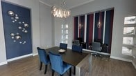 This single-floor space makes a strong design statement through its thoughtful combination of bold aesthetics and refined details. The centerpiece of the main room is a large dark dining or conference table surrounded by plush navy blue velvet chairs, offering both comfort and style. Above, a striking cluster chandelier with globe-shaped glass pendants draws the eye upward, highlighting the room's generous ceiling height.

The accent wall serves as a dramatic focal point, featuring bold vertical stripes in deep navy and burgundy against a white background, adding depth and a sense of sophistication to the space. A large mirror on the accent wall reflects the room, enhancing the sense of openness and amplifying the natural light that filters in through a tall, multi-pane window nearby.

The flooring throughout is a warm wood-tone laminate or hardwood, grounding the cool color palette and adding an inviting contrast to the grey walls. Crown molding along the upper edges of the walls reinforces the space's polished, upscale character. Additional pendant lighting in the background hints at further zones within the space, suggesting a versatile layout suited for meetings, presentations, or collaborative work. Overall, the space exudes a confident, modern elegance.