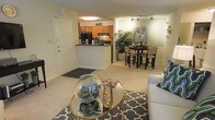 This single-floor residence showcases an open-concept design that seamlessly integrates the living, dining, and kitchen areas into one flowing space. The living area is anchored by plush carpeting and a stylish glass-topped coffee table with gold accents, complemented by a patterned area rug that adds visual interest to the room. A comfortable sofa and tasteful decorative elements, including a glass terrarium centerpiece, contribute to the welcoming atmosphere.

The kitchen features warm honey-toned cabinetry, a dark countertop peninsula that provides additional workspace and serves as a natural divider between the kitchen and living areas, and black appliances. Track lighting illuminates the kitchen and dining zone effectively. The dining area accommodates a dark high-top table with matching woven chairs, creating a casual yet stylish setting for meals. Lush indoor plants positioned near the dining area add a refreshing natural element throughout the space.

The interior is finished with neutral, warm-toned walls and consistent carpeting, creating a cohesive and inviting ambiance throughout the residence.