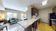 This single-floor unit offers an open-concept layout that seamlessly integrates the kitchen, living, and sleeping areas into one cohesive space. Light hardwood flooring runs throughout, lending warmth and visual continuity across the unit.

The kitchen is well-appointed with rich dark-wood cabinetry extending floor to ceiling, providing generous storage. Stainless steel appliances — including a range, microwave, and refrigerator — are complemented by a marble-patterned countertop that wraps around a peninsula breakfast bar. The bar accommodates seating for two and serves as a natural divider between the kitchen and the main living area. A pendant light fixture above the peninsula adds a refined touch.

The living area is generously proportioned, featuring a large window that fills the room with natural light and offers views of the surrounding cityscape. The sleeping area, open to the living space, benefits from multiple windows that further enhance brightness and the connection to the outdoors.

The bathroom is finished with large-format tile flooring, a floating dark-wood vanity with an undermount sink, and a full-width mirror. The overall aesthetic throughout the unit is contemporary and cohesive, with warm neutral tones and consistent design elements tying each area together.