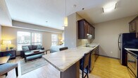 This single-floor unit offers an open-concept layout that seamlessly integrates the kitchen, living, and sleeping areas into one cohesive space. Light hardwood flooring runs throughout, lending warmth and visual continuity across the unit.

The kitchen is well-appointed with rich dark-wood cabinetry extending floor to ceiling, providing generous storage. Stainless steel appliances — including a range, microwave, and refrigerator — are complemented by a marble-patterned countertop that wraps around a peninsula breakfast bar. The bar accommodates seating for two and serves as a natural divider between the kitchen and the main living area. A pendant light fixture above the peninsula adds a refined touch.

The living area is generously proportioned, featuring a large window that fills the room with natural light and offers views of the surrounding cityscape. The sleeping area, open to the living space, benefits from multiple windows that further enhance brightness and the connection to the outdoors.

The bathroom is finished with large-format tile flooring, a floating dark-wood vanity with an undermount sink, and a full-width mirror. The overall aesthetic throughout the unit is contemporary and cohesive, with warm neutral tones and consistent design elements tying each area together.