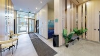 This contemporary building lobby offers a polished and functional entry experience, thoughtfully designed with modern aesthetics and practical amenities. The space features large-format stone-look tile flooring throughout, complemented by recessed pot lighting and linear LED strip lighting that create a bright, welcoming ambiance. Natural light filters in through the full-height glazed entrance doors, enhancing the open feel of the corridor.

The lobby is defined by distinctive light wood vertical columns and paneling that add warmth and visual interest, contrasting elegantly with the neutral grey tones of the walls and floors. A dedicated seating area with modern upholstered chairs and round side tables provides a comfortable waiting space for residents and guests. Tall decorative planters with lush greenery add a fresh, organic element to the interior.

A stainless steel passenger elevator serves as a central focal point at the rear of the lobby, flanked by large-format stone tile wall cladding. Parcel lockers are conveniently located near the entrance, offering a practical package management solution. The lobby also includes a hand sanitizer station and clearly marked accessible doorways, reflecting a commitment to accessibility and resident well-being. Overall, the space strikes a balance between modern design and everyday functionality.