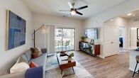 Located at 9911 Dessau Rd in Austin, TX, this single-story unit offers a thoughtfully designed open-concept layout that seamlessly connects the living, dining, and kitchen areas. The living room is bright and welcoming, featuring wood-look plank flooring, a ceiling fan, and large sliding glass doors that open onto a private balcony with outdoor seating and views of the surrounding community. An elegant arched passageway connects the living space to the kitchen, which is outfitted with dark wood cabinetry, granite countertops, subway tile backsplash, and stainless steel appliances including a range, microwave, and refrigerator. A kitchen island with bar seating provides additional prep and dining space. Adjacent to the kitchen, a dedicated dining nook accommodates a round table and chairs, offering a cozy spot for meals. The bedroom features plush carpet flooring, a ceiling fan, natural light from a window, and ample wall space. The full bathroom includes a granite-topped vanity with dark wood cabinetry, a tub-shower combination with tile surround, and wood-look flooring. Clean white walls and consistent contemporary finishes throughout the unit create a cohesive and modern aesthetic.