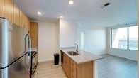 This single-floor space showcases a clean, modern design centered around an open-concept layout that seamlessly connects the kitchen and living areas. The kitchen is anchored by a peninsula-style island topped with speckled granite countertops, providing both additional workspace and a casual dining or entertaining surface. Warm-toned wood cabinetry with sleek bar-pull hardware offers generous storage, complemented by an integrated stainless steel sink with a contemporary arc faucet and a built-in dishwasher. Recessed lighting is thoughtfully positioned throughout the ceiling, ensuring consistent, even illumination across the space. The light hardwood-style flooring extends uniformly from the kitchen into the adjoining living area, contributing to a cohesive and airy atmosphere. Crisp white walls enhance the sense of openness, while a window in the living area allows natural light to filter in, brightening the interior. The layout flows naturally toward additional rooms visible in the background, suggesting practical connectivity between spaces. Overall, the unit presents a well-appointed, contemporary living environment with quality finishes and a functional, open design aesthetic.