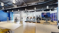 Located at 1100 East Apache Boulevard in Tempe, Arizona, this fitness center offers a well-appointed, single-floor workout facility spanning a generous footprint designed to accommodate a full range of exercise activities. The space is characterized by its industrial-modern aesthetic, featuring exposed ductwork and piping along the dark ceiling, which contrasts with bright white walls and a bold blue accent wall that anchors the strength training zone. Flooring transitions between light hardwood-style surfaces in the cardio areas and durable speckled rubber flooring in the free weights and strength equipment sections, providing both functionality and visual definition throughout the space. The cardio zone is equipped with multiple treadmills, ellipticals, rowing machines, and stair climbers, accompanied by a large display screen. The strength area includes cable machines, a Smith machine, and a rack system with accessories. A row of floor-to-ceiling glass garage doors lines two sides of the facility, flooding the interior with abundant natural light and offering direct views of an outdoor pool and courtyard area. Pendant lighting and track fixtures supplement the natural light. A reception counter with seating is positioned near the entrance, and large mirrors in the strength zone enhance the sense of openness throughout this energetic, well-equipped fitness space.