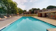 Located at 3822 Pembrook Ct in Columbus, Georgia, this outdoor community amenity area centers around a generously sized swimming pool with clear, blue-green water. The pool deck is finished in concrete and provides ample space for relaxation, lined with orange lounge chairs positioned for sunbathing. Shaded seating options are available via large orange market umbrellas paired with small tables and chairs, offering a comfortable retreat from the sun.

The entire pool area is enclosed by a chain-link fence, providing a secure environment with a gated entry point adjacent to a brick building. The fencing ensures a defined boundary while maintaining open sightlines to the surrounding mature tree canopy, which provides a natural, green backdrop. A lamp post stands within the enclosure, suggesting the space is usable during evening hours as well.

Beyond the pool deck, a grassy area is visible through the fence, along with what appears to be an outdoor grilling or gathering structure, extending the recreational possibilities of the space. The surrounding residential buildings, constructed in brick, are visible in the background, reinforcing the community-oriented nature of this amenity. Overall, the space presents a well-organized, functional outdoor recreational area suited for warm-weather leisure.