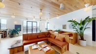 This single-floor space combines thoughtful design with versatile functionality across several distinct zones. The main lounge area is anchored by custom-built natural wood sectional seating with terracotta-toned cushions, complemented by woven rattan coffee tables and integrated booth-style seating partitions that divide the space into intimate gathering areas. A warm wood-planked ceiling overhead adds texture and coziness, while globe and cone pendant fixtures provide ambient lighting throughout. Floor-to-ceiling windows along the perimeter flood the interior with natural light and offer panoramic city rooftop views.

A secondary lounge features a large sage green sectional sofa with coordinating cushions and woven cube ottomans, connected to the main area through an open doorway. The kitchenette is well-appointed with white cabinetry, a light countertop, terracotta zellige tile backsplash, open shelving stocked with dishware, a deep sink, wine refrigerator, and under-counter appliances, all accented by brushed gold wall sconces.

A media room provides a large wall-mounted television, light wood console, and French doors opening to a private outdoor terrace with city views. Throughout, lush tropical plants, curated artwork, and light-toned flooring contribute to a cohesive, warm, and welcoming ambiance.