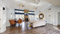 Located at 100 Arbor Lake Boulevard in Hermitage, Tennessee, this single-floor community living room is a standout gathering space defined by its dramatic vaulted ceilings and open, airy layout. The soaring roofline is accented by clerestory windows that flood the interior with natural light, complemented by dual ceiling fans with gold-toned fixtures that add a warm, polished touch overhead.

A floor-to-ceiling stacked stone fireplace serves as the architectural centerpiece of the room, anchoring a bold navy accent stripe that runs horizontally across the upper walls, creating a striking visual contrast against the bright white surfaces. Rich hardwood-style flooring flows throughout, grounding the space with warmth and texture.

The layout accommodates multiple functional zones, including a comfortable lounge area with a tufted sofa, colorful accent pillows, and a patterned area rug, as well as a workstation area with a desk and seating. A decorative circular wood-framed mirror adds an artistic element to the lounge side of the room.

Large glass-paned French doors on multiple walls open the space to outdoor views and provide seamless indoor-outdoor connectivity. A bright, glass-windowed entry vestibule with a tufted bench offers a welcoming transition into the space. Together, these elements create a functional, stylish, and inviting community environment.