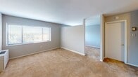 This single-floor residential unit offers a straightforward and functional layout designed for comfortable everyday living. The main living area is generously sized, featuring wall-to-wall beige carpeting and neutral gray walls that create a clean, versatile backdrop. A large window fitted with horizontal blinds allows natural light to fill the space, contributing to a bright and open atmosphere. An open passageway connects the living area to an adjacent room, painted in a soft blue-gray tone, providing a sense of continuity throughout the unit. One wall in the living area features a distinctive grid-patterned tile surface, adding a subtle textural element to the otherwise understated interior. The kitchen is compact yet well-equipped, offering light wood cabinetry, a gas range, a refrigerator, and a stainless steel sink. A built-in breakfast bar with a laminate countertop provides a convenient casual dining option. The kitchen flooring consists of neutral tile that contrasts with the carpeted living spaces. The bathroom is efficiently designed, featuring a combined tub and shower, a toilet, and tile flooring. Overall, the unit presents a clean, move-in ready environment with a practical and cohesive design throughout.