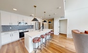This contemporary two-story residence offers 1,740 square feet of thoughtfully designed living space, with approximately 870 square feet on each floor. The open-concept kitchen serves as a central highlight, featuring white shaker cabinetry, a large quartz waterfall island with an undermount sink, stainless steel appliances including a French door refrigerator and range, a textured tile backsplash, and striking matte black pendant lights with brass accents — complemented by plaid upholstered bar stools that add a pop of color. Warm hardwood-style flooring runs throughout both levels, enhancing the bright, clean aesthetic. Multiple bedrooms are designed for functionality, each featuring a platform bed with built-in storage, a dedicated desk workspace, and large windows that frame urban cityscape views. Exposed concrete ceilings and structural columns lend an industrial character to select rooms. The upper floor includes a landing with an open black metal railing and expansive windows overlooking the surrounding city. Multiple bathrooms are finished with backlit LED mirrors, light wood vanities with quartz countertops, walk-in glass-enclosed showers with matte black fixtures, and hardwood-style flooring — delivering a clean, modern aesthetic throughout.