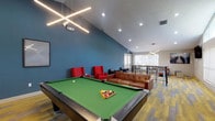 This single-floor amenity lounge is designed for recreation and social gatherings, combining vibrant design elements with versatile furnishings. The space is anchored by a full-size billiards table with a green felt surface, positioned beneath a striking cross-shaped linear pendant light fixture that serves as a bold architectural focal point. Soaring vaulted ceilings amplify the sense of openness throughout the room.

The lounge is divided into distinct seating zones. A rich cognac leather sofa and a pair of vivid red accent chairs create a casual conversation area near the pool table, while a separate lounge area offers additional seating with grey upholstered chairs arranged around a communal table. A wall-mounted flat-screen television is positioned for comfortable viewing from the seating areas.

The color palette features a deep teal accent wall that contrasts with the light grey walls and white ceiling, adding depth and visual interest. Patterned carpet tiles in yellow, grey, and brown tones run throughout the space, complementing the overall contemporary aesthetic. Decorative wall art enhances the interior design.

Large windows fitted with sheer roller shades flood the room with natural light and offer views of an outdoor pool area and surrounding grounds. A set of white-framed French doors provides direct access to the exterior, further connecting the indoor space to the outdoor amenities.