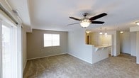 This single-floor space is designed with an open-concept layout that promotes a sense of airiness and functionality. The living area is finished with wall-to-wall carpet in a neutral beige tone, providing a comfortable and cohesive foundation throughout the space. A multi-blade ceiling fan with an integrated light fixture is centrally mounted, offering both practical ventilation and ambient lighting. The walls are painted in a light neutral tone, complementing the bright, clean aesthetic of the space.

A window on the far wall allows natural light to filter into the living area, enhancing the overall brightness of the room. The living space transitions smoothly into the kitchen area via a raised breakfast bar with a light-colored countertop, ideal for casual dining or additional workspace. The bar features an overhang that accommodates seating. Above the kitchen area, pendant lights hang at varying heights, adding a layered lighting element. A mirrored surface near the kitchen entry reflects additional light, further contributing to the open and welcoming atmosphere of this well-designed space.