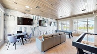 Located at 1708 Starship Dr in Cocoa, Florida, this well-appointed game room offers a dynamic and inviting atmosphere designed for leisure and social interaction. The single-floor space is thoughtfully laid out to accommodate a variety of recreational activities without feeling cramped.

The room's most striking architectural feature is its wood-planked ceiling, which adds warmth and a contemporary rustic charm. Herringbone-patterned flooring runs throughout, lending a refined texture underfoot. A statement accent wall clad in reclaimed-style wood planks anchors one side of the room, complete with built-in open shelving displaying décor and books, alongside a wall-mounted flat-screen television.

On the opposite end, a bold artistic mural wall in abstract, typographic style serves as a dramatic backdrop for dual wall-mounted TVs and a casual dining table with upholstered chairs — ideal for game nights or casual gatherings. A sleek pool table takes center stage beneath a modern multi-bulb pendant chandelier, while a shuffleboard table offers additional entertainment options nearby.

Plush lounge seating and area rugs create comfortable conversation zones throughout. Floor-to-ceiling windows and glass doors flood the space with natural light and frame views of the outdoor pool area and surrounding greenery, seamlessly connecting the interior to the Florida landscape beyond.