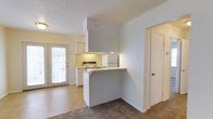 This single-floor space is designed with an open layout that connects the living area to the kitchen, creating a functional and cohesive flow throughout. The kitchen is equipped with white cabinetry, a gas range, and a refrigerator, all complemented by under-cabinet lighting that brightens the workspace. A peninsula-style breakfast bar serves as a natural divider between the kitchen and the adjacent living area, offering additional counter space and casual seating potential. The kitchen and entry zone feature tile flooring, while the living area transitions to carpet, subtly defining each functional zone. A textured popcorn ceiling runs throughout, and the walls are painted in a neutral, light tone that keeps the space feeling bright and open. A paneled door at the rear of the kitchen floods the space with natural light and suggests access to an outdoor area. A secondary door is also visible along the adjacent wall, providing additional access or storage. Overall, the space maintains a clean, practical aesthetic suited for everyday living.