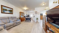 This thoughtfully designed single-floor home offers a seamless, open-concept layout that connects the living, dining, and kitchen areas into one cohesive living space. Light-toned wood-style flooring flows throughout the main areas, complemented by neutral white walls that create a bright and welcoming atmosphere. The living area features a comfortable seating arrangement centered around a fireplace, with windows that bring in natural light and views of the surrounding neighborhood. A dedicated dining space with a dark-toned table and chairs sits adjacent to the kitchen, which is designed in a classic galley style with white cabinetry, dark countertops, stainless steel appliances including a refrigerator and dishwasher, and a back door that provides additional access and natural light. The primary bedroom is generously sized, featuring rich wood furnishings and a large window with outdoor views. A second bedroom offers a clean, functional space with light wood flooring and a bright window. The full bathroom is finished with white cabinetry and dark countertops, and includes a shower-tub combination. Conveniently located off the bathroom is a dedicated laundry area equipped with a full-size washer and dryer. Overall, the home presents a functional, comfortable layout with a warm and inviting character throughout.