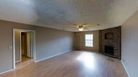 This single-floor space offers a comfortable and functional layout highlighted by several classic architectural details. The main living area is generously sized, featuring smooth hardwood-style flooring that reflects natural light pouring in through a double-hung window, creating a bright and welcoming atmosphere. A ceiling fan with a built-in light fixture is centrally positioned, providing both air circulation and ambient lighting. A standout feature of the room is the floor-to-ceiling brick fireplace situated in the corner, adding warmth and a timeless, traditional character to the space. The walls are finished in a neutral warm gray tone, complementing the light-toned flooring and contributing to a cohesive, understated aesthetic. The textured popcorn ceiling runs throughout, adding a layer of visual depth overhead. A doorway and an additional interior door provide access to adjoining spaces, suggesting a practical and connected floor plan. White baseboards frame the perimeter of the room cleanly, offering a crisp contrast to the neutral walls. Overall, the space balances classic design elements with an open, airy feel well-suited for everyday living.